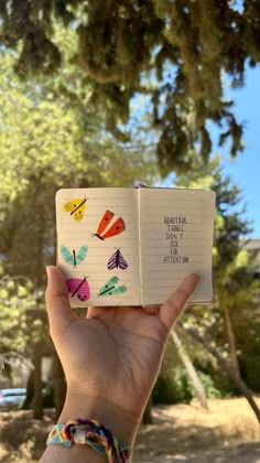 a hand holding an open notebook with colorful butterflies on it, in front of a tree