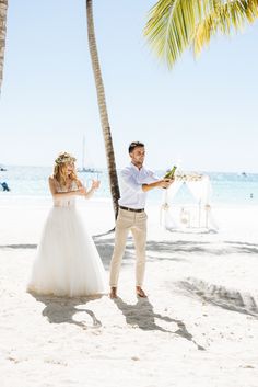 a man and woman standing on top of a sandy beach next to the ocean holding wine glasses