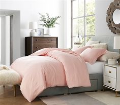 a bed with pink comforter and pillows in a white room next to a window