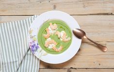 a white plate topped with green soup and shrimp on top of a wooden table next to a spoon