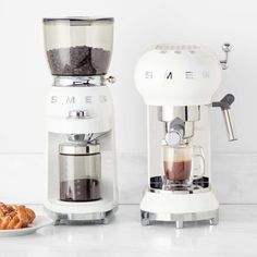 two coffee makers sitting next to each other on top of a white counter with pastries in front of them