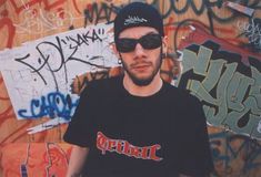 a man wearing sunglasses and a black shirt standing in front of graffiti covered wall