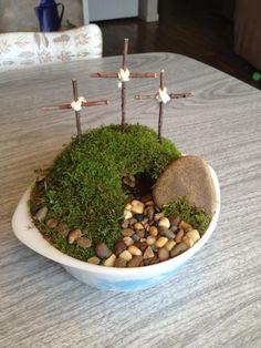 a white bowl filled with rocks and grass on top of a wooden table next to a cross