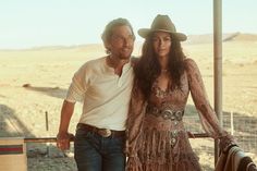 a man and woman standing next to each other in front of a fence with an open field behind them