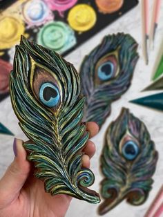 a hand holding a peacock feather broochle in front of some paintbrushes