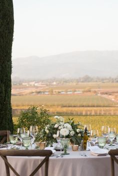 the table is set with white flowers and wine glasses for an outdoor dinner in front of a vineyard