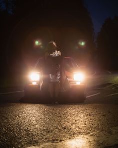 a woman standing on the hood of a car in the dark with headlights shining down