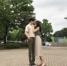 a young man and woman standing next to each other on a brick walkway in front of trees