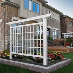 a white trellis in front of a brick house with flowers on the lawn and landscaping around it