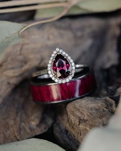 A matching wedding ring set from Staghead Designs sits on a piece of wood surrounded by leaves. On top sits a 14K white gold engagement ring with a pear cut garnet center stone & diamond halo. Below sits a black zirconium wedding band in our "Rainier" single-channel design with purpleheart wood inlay. Wedding Garnet Solitaire Rings, Luxury Garnet Engagement Ring, Elegant Garnet Diamond Ring, Garnet Halo Engagement Ring, Red Garnet Ring With Halo Setting, Ruby Wedding Ring Set, Matching Wedding Ring Sets, Matching Ring Set, Salt Pepper Diamond