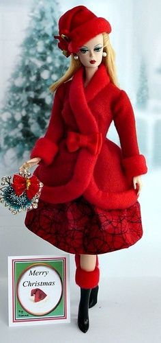 a doll dressed in red is standing next to a christmas card and a small tree