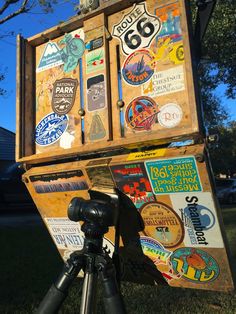 an old wooden box with stickers on it sitting in the grass next to a tripod