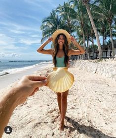 a woman is standing on the beach wearing a hat and holding her hand up to her head