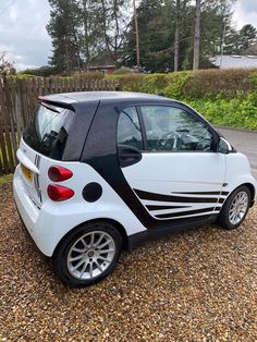 a small smart car parked on gravel next to a wooden fence