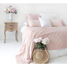 a bed with pink linens and pillows in a white room next to a table with flowers