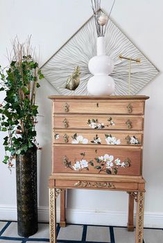 an old dresser with flowers painted on it next to a potted plant and mirror