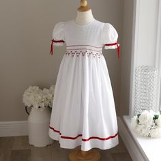 This Strasburg Smocked Heirloom White Dress W/ Red Trim Is A Classic! One Of The Original Strasburg's Most Popular Designs, This Sweet Dress Has Lots Of Detail. The Bodice Is Fully Smocked With A Small Lattice Design With A Red Horizontal Band Accent With Accordion Smocking Just Above The Waist, Followed By The Same Lattice Detail. It Is Finished Off With Lovely Red And White Hand-Embroidered Rosettes With Green Leaves In A Red Embroidered Chevron Pattern. The Puff Sleeves Gather With A Red Sati Smocked Christmas Dresses, Smocking Patterns, English Wedding, Lattice Design, Popular Designs, White Hand, Smocked Dress, Fabric Covered Button, Red Satin