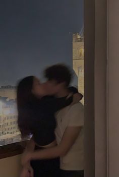 a man and woman kissing in front of a window with the city lights behind them