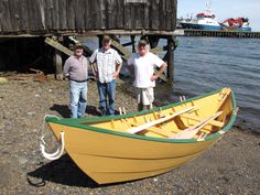 three men standing next to a yellow and green boat on the shore with a dock in the background