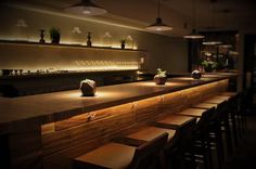 an empty bar with wooden stools and lights on the wall above it, along with several potted plants