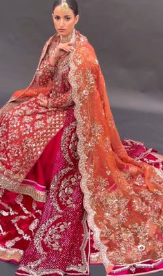 a woman in a red and gold bridal dress sitting on a gray background with her hands clasped to her chest