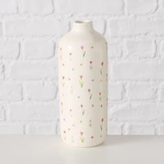 a white vase sitting on top of a table next to a brick wall with flowers painted on it