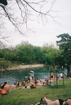 many people are sitting on the grass by the water and in the park, one person is laying down