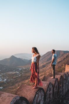 two people are standing on the edge of a wall looking out at mountains and valleys