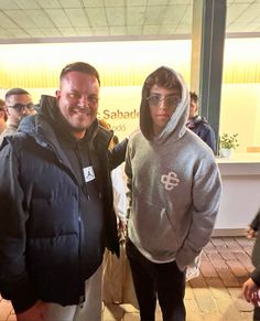 two men standing next to each other in front of a store with people looking at the camera