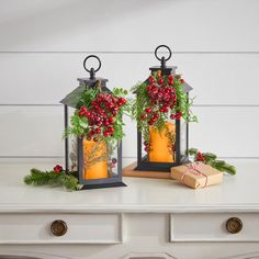 two lanterns decorated with holly and red berries sit on a white table next to a present