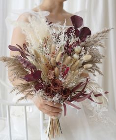 a bride holding a bouquet of dried flowers and feathers in her hands, with white curtains behind her