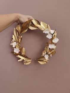 a hand holding a gold and white wreath with flowers on the front, against a pink wall