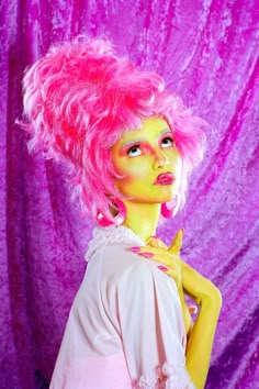 a woman with pink hair is posing in front of a purple background and wearing bright makeup