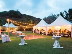 a group of people standing around tables and chairs under white tents with lights on them