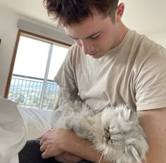 a man holding a cat in his arms while sitting on a bed next to a window