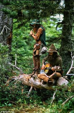 two men are sitting on a log in the woods playing guitar and singing while another man stands next to them