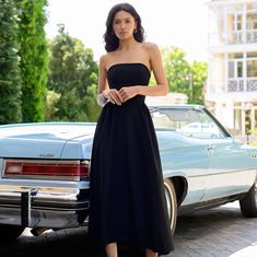 a woman standing in front of a car wearing a strapless black dress and heels