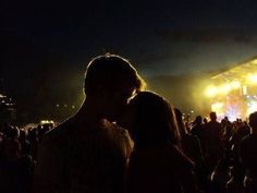 a couple kissing in front of a crowd at a music festival with bright lights on the stage behind them