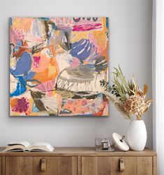 an abstract painting hangs on the wall next to a wooden cabinet and vase with flowers