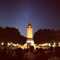 a crowd of people standing in front of a tall clock tower at night with lights on