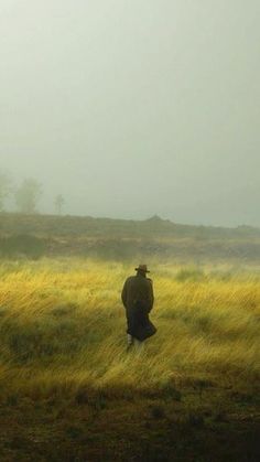 a man in a hat and coat is walking through the grass on a foggy day