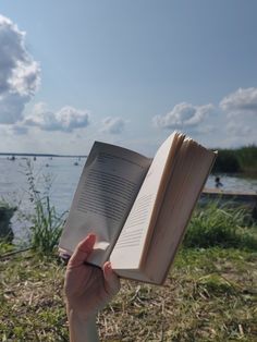 a person is holding an open book in their hand near the water with boats on it