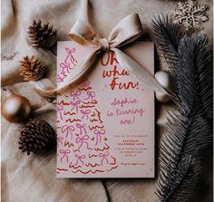 a pink and red christmas party card on top of a table with pine cones, ornaments and ribbon