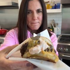 a woman holding up a large sandwich with toppings on it in her hands and looking at the camera