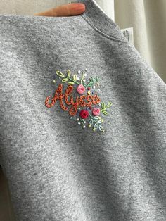 someone is holding up a gray sweatshirt with an embroidered name and flowers on the front