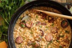 a pot filled with beans and sausages on top of a wooden spoon next to carrots