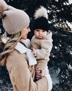 a woman holding a baby wearing a hat and coat in front of a pine tree