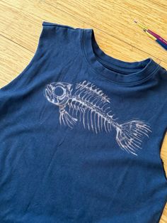 a t - shirt with a fish skeleton on it sitting on the floor next to crayons