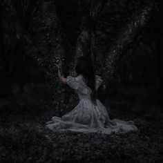 a woman in a white dress sitting on the ground under a tree with her hands out