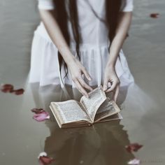 a woman kneeling down reading a book on the ground with rose petals around her feet